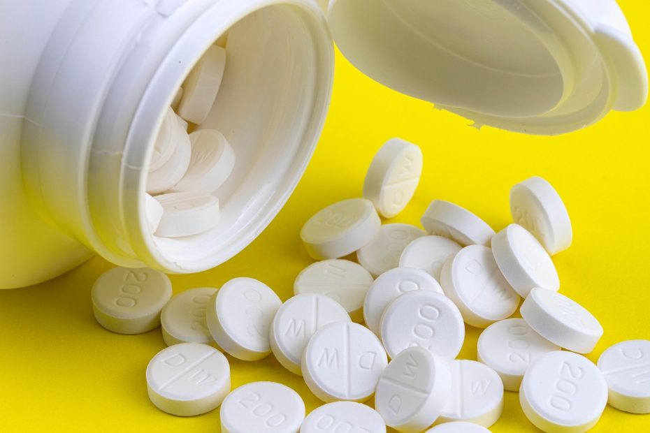 Białe tabletki farmaceutyczne wysypane z otwartego pojemnika na żółtym tle.